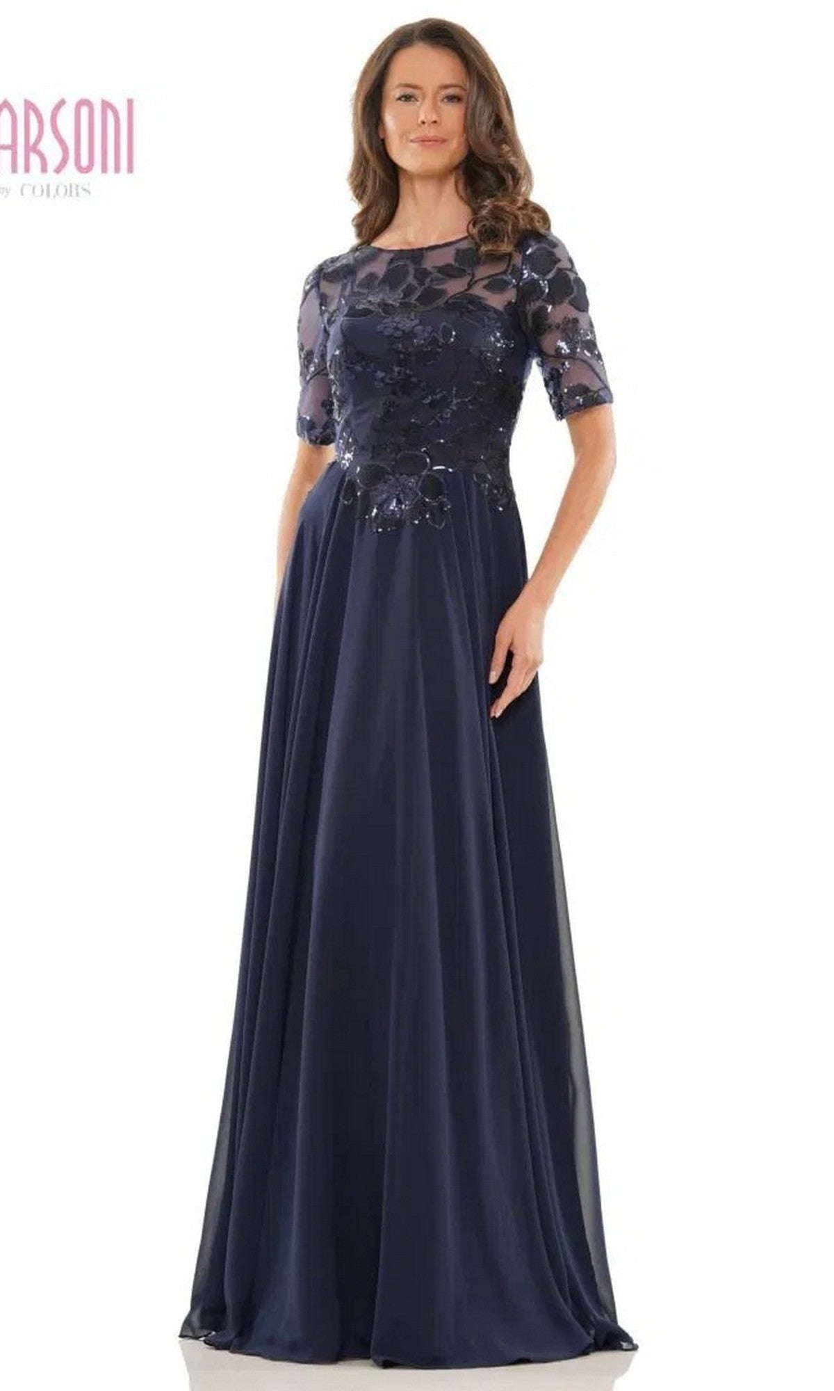A woman models the Marsoni M286 long formal dress, a floor-length navy gown with sheer floral embroidered sleeves and bodice, standing against a white background.